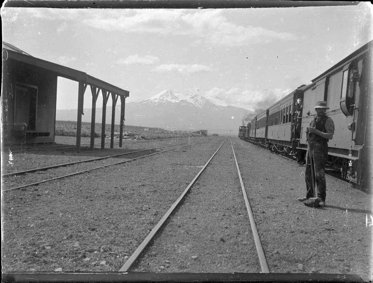 View along a train stopped at Waiouru Railway Station, railway track in the centre, and the corner of a station building on the left. Mount Ruapehu is in the background, and a railway employee is standing by the train wi